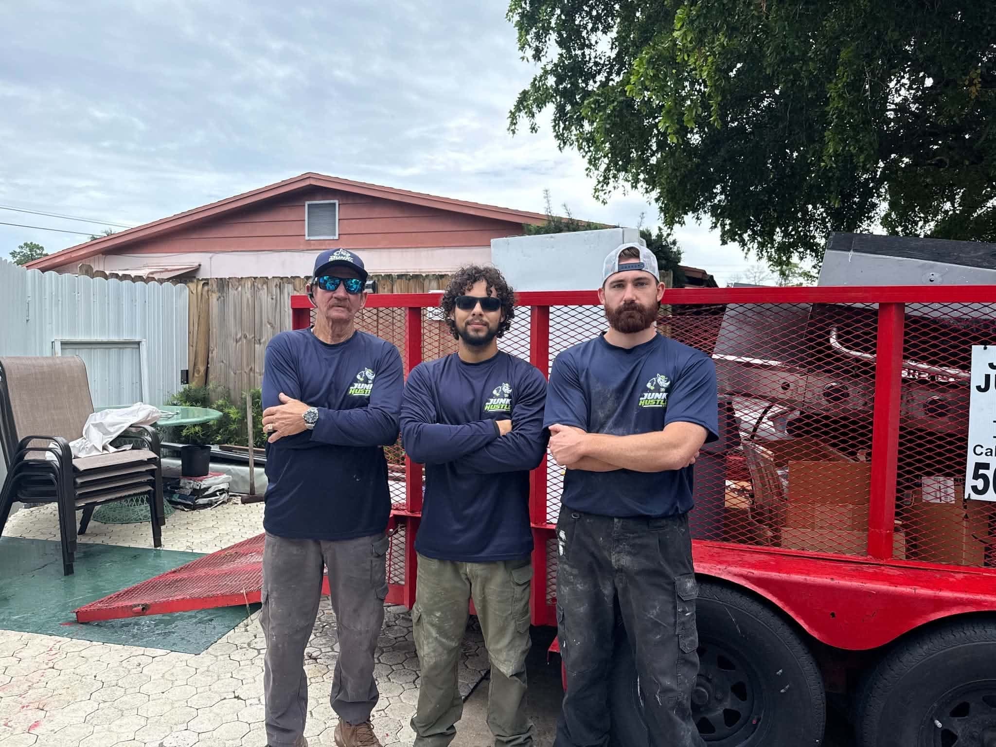 The JunkHustle crew in navy JunkHustle shirts, arms crossed, in front of the red trailer