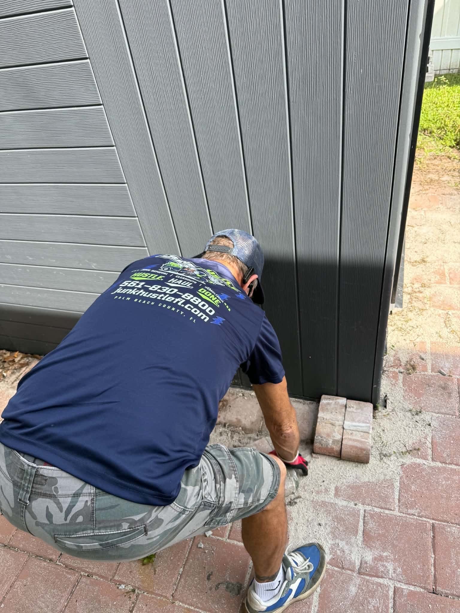 A JunkHustle crew member working at the base of a large gray resin shed on a paver patio